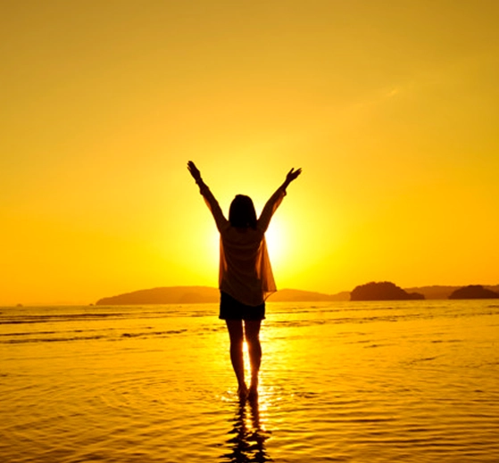 person with arms raised standing in the water with a golden hue from the sun