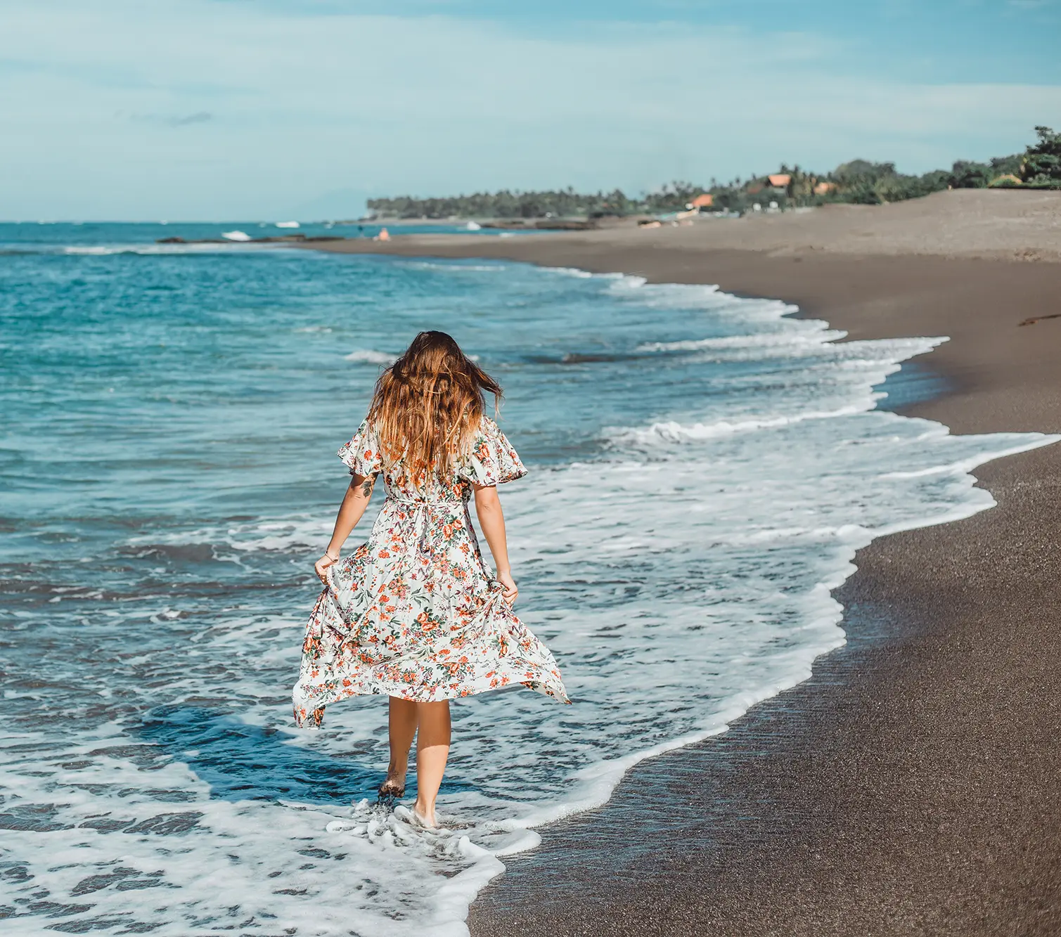 woman in flowered dress walking along the beach next to the sand